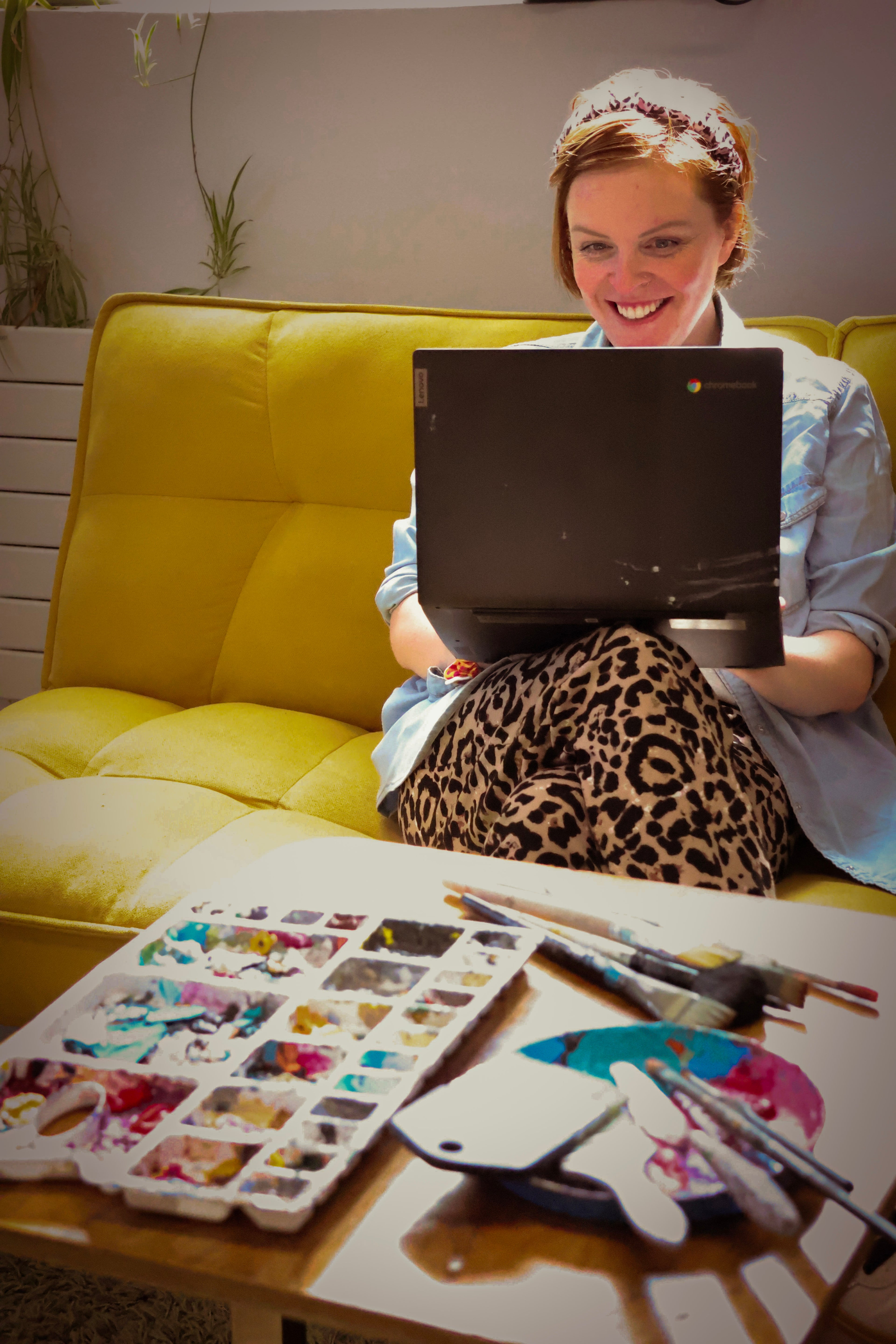 Person sitting on a yellow couch with a laptop, surrounded by art supplies on a table.
